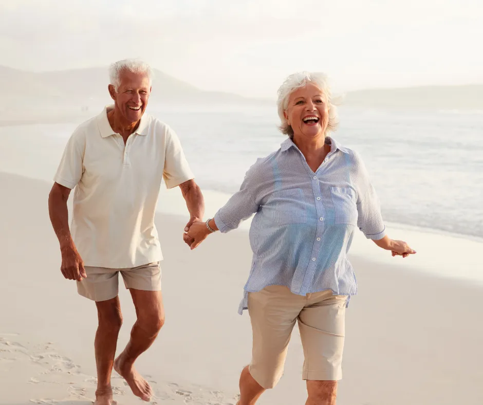 Happy retired couple holding hands and walking along a sandy beach, enjoying a stress-free retirement.