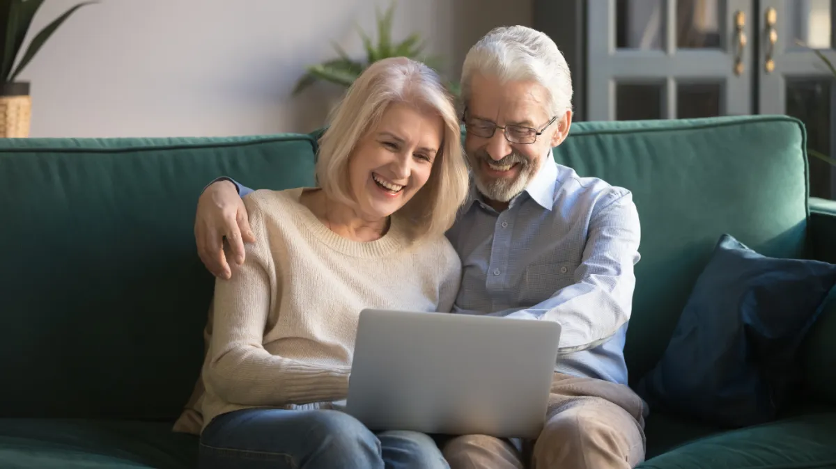 Smiling older couple sitting together on a green couch, looking at a laptop screen while planning for retirement.