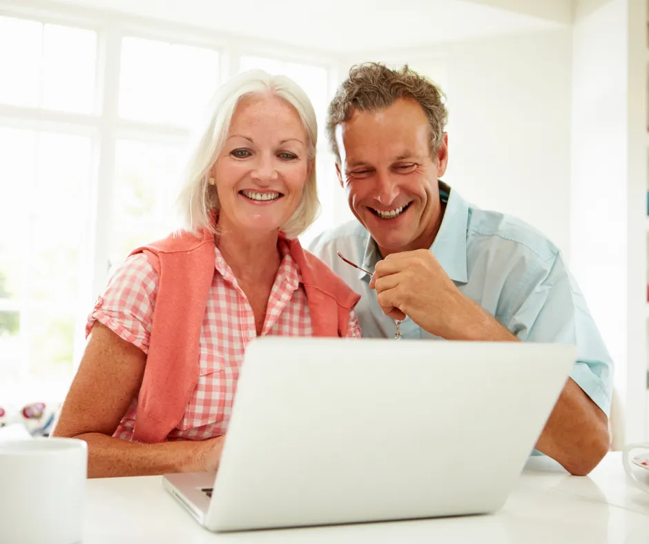 Smiling middle-aged couple sitting together at a desk, looking at a laptop screen in a bright, modern home setting.