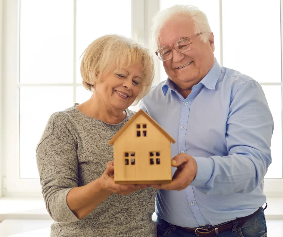 Smiling older couple holding a small wooden house model, symbolizing financial security, home ownership, and planning for their future.