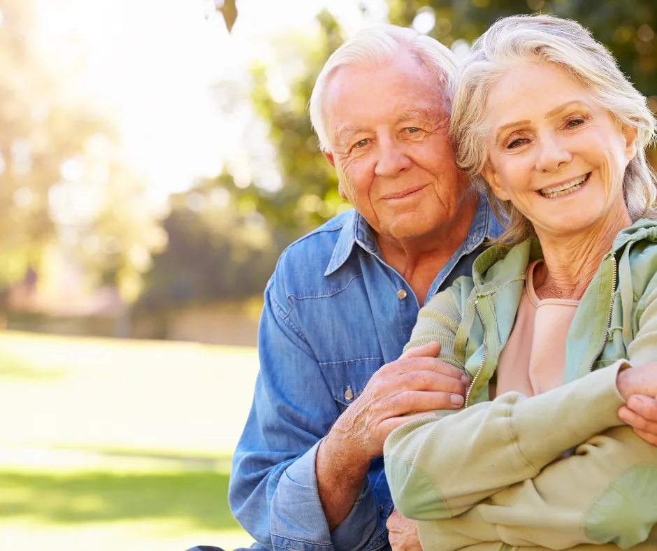 Happy senior couple sitting together outdoors, smiling at the camera, with a sunny park background. The woman is wearing a green hoodie, and the man is wearing a denim shirt, symbolizing a relaxed and secure retirement.