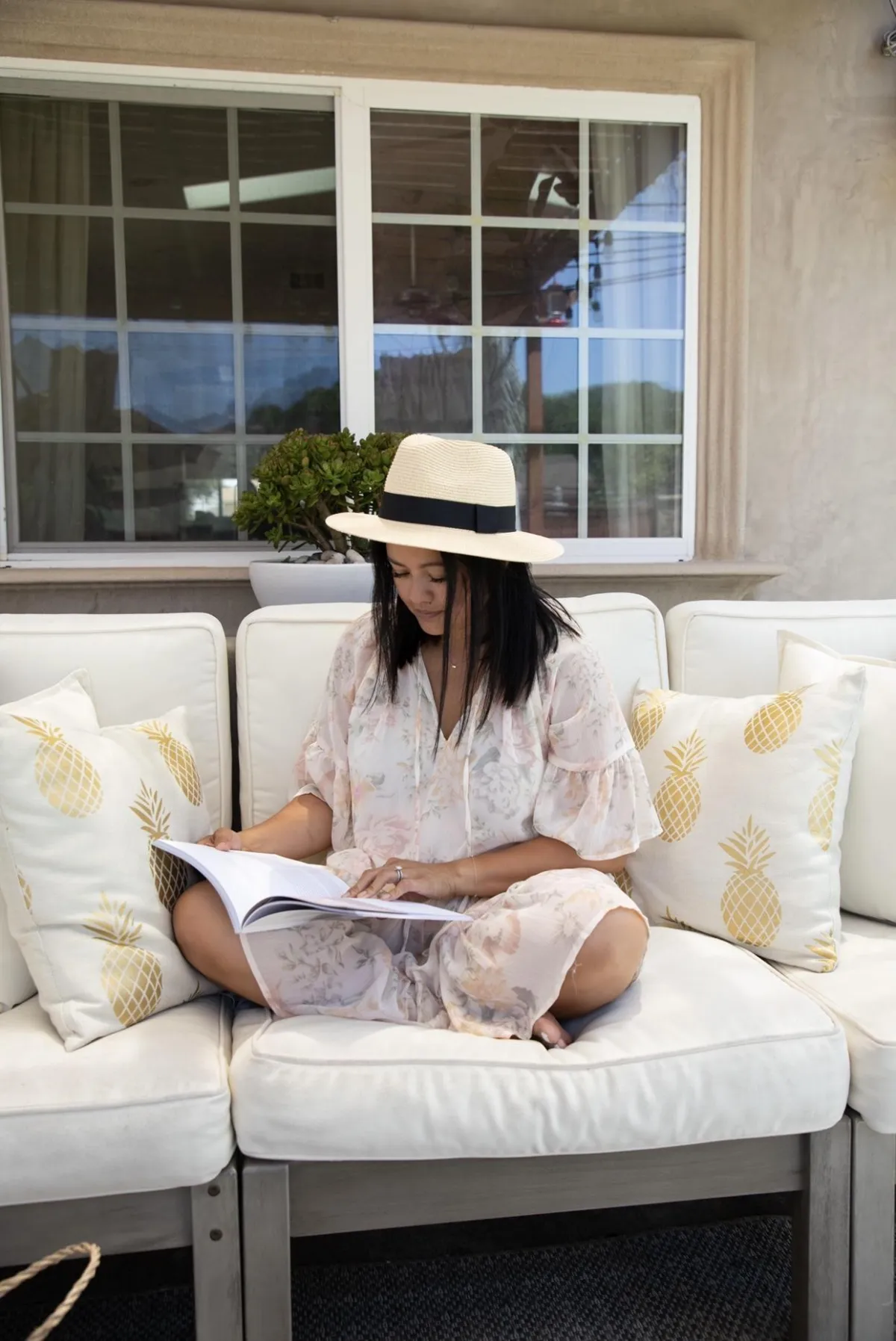 Childless woman in a floral dress and straw hat reading a book on a patio sofa with pineapple-print pillows.