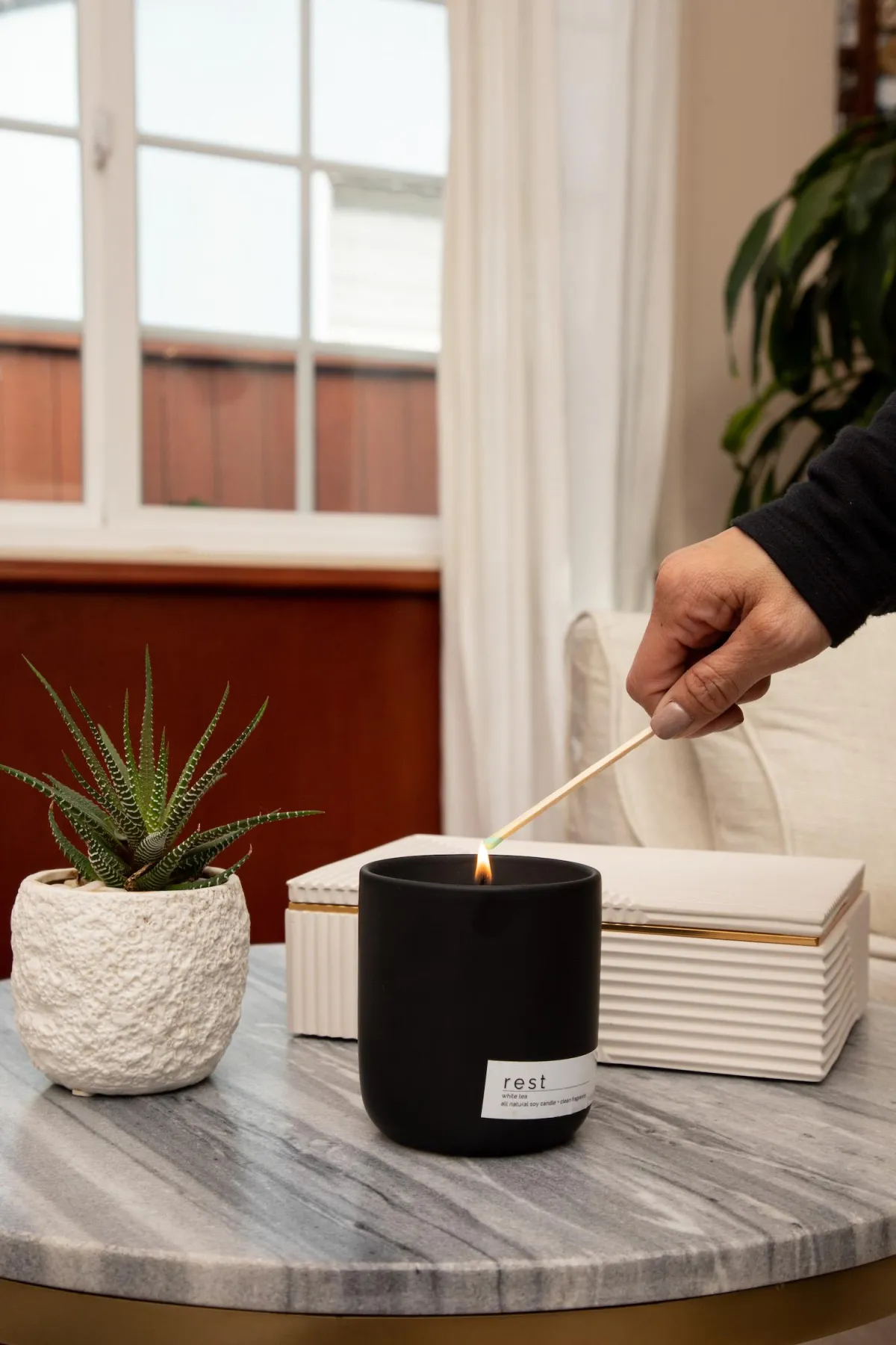 Hand lighting a black candle labeled “rest” on a marble table beside a small succulent and ceramic box in a cozy living room. childless not by choice