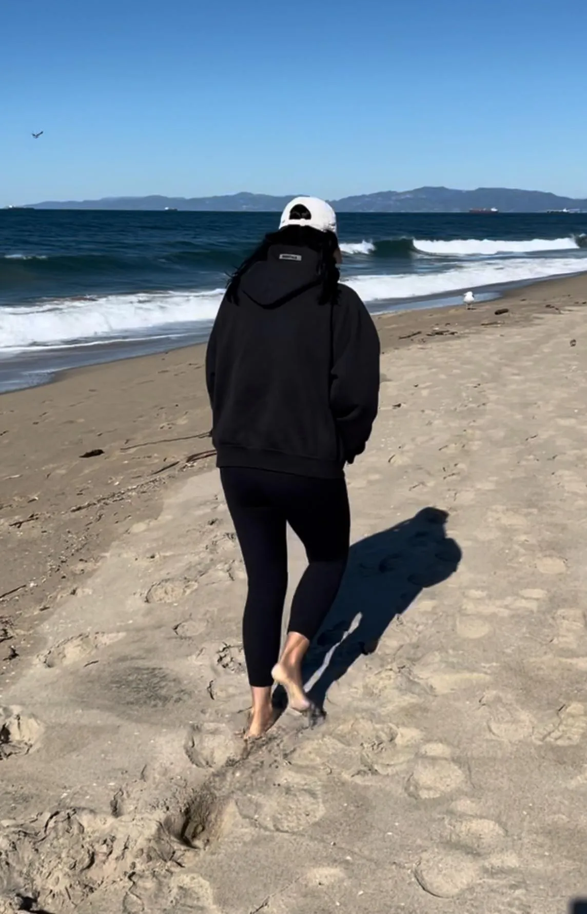 Childless Woman walking barefoot on a sandy beach in a black hoodie and leggings, facing the ocean waves.