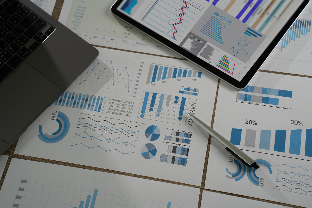 Hands holding a pen over printed financial charts and graphs on a wooden desk, with a laptop showing analytics software in the background