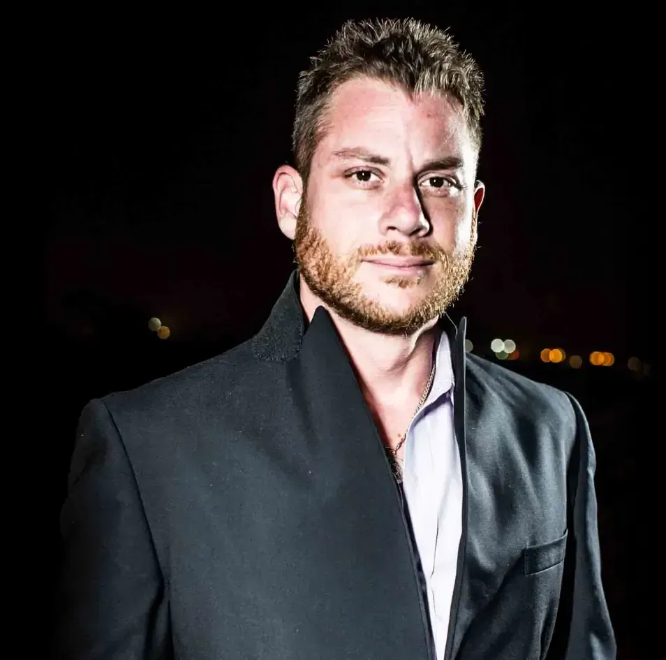 Official picture of Brandon Mushlin: A bearded man with short hair stands in a light-colored shirt and dark jacket, in front of a dark background with blurry lights.