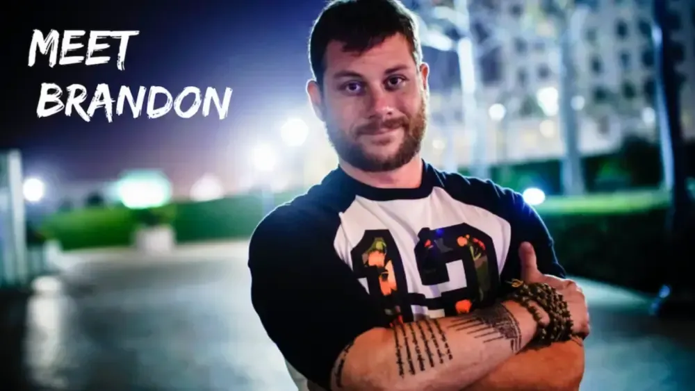 A man stands confidently with arms crossed. The text "Meet Brandon Mushlin" appears on the left. The background shows a blurred outdoor night scene with lights and buildings.