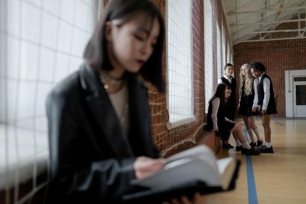 A young girl sits alone at school, while a group of girls in the background point and whisper about her