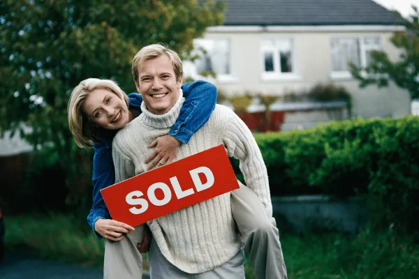 Happy Homeowners in Front of a “For Sale” Sign