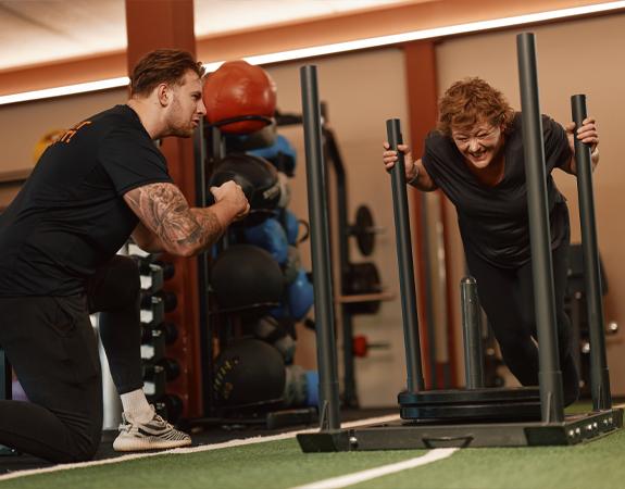A smiling personal trainer standing next to a seated client using a piece of gym equipment. Warm indoor lighting.