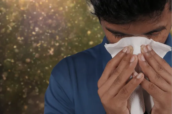 Person sneezing into a tissue with blurred pollen in the background, representing seasonal allergies.
