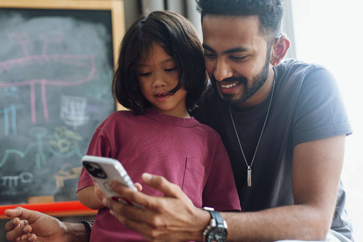 young adult with a hearing aid watching phone with his daughter