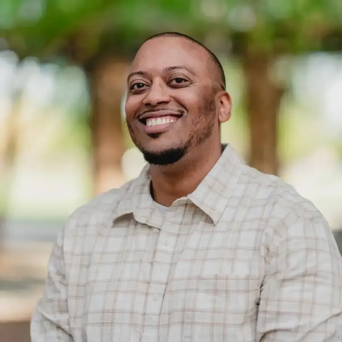 Damian Coln smiling in a beige plaid shirt standing outdoors with trees blurred in the background.