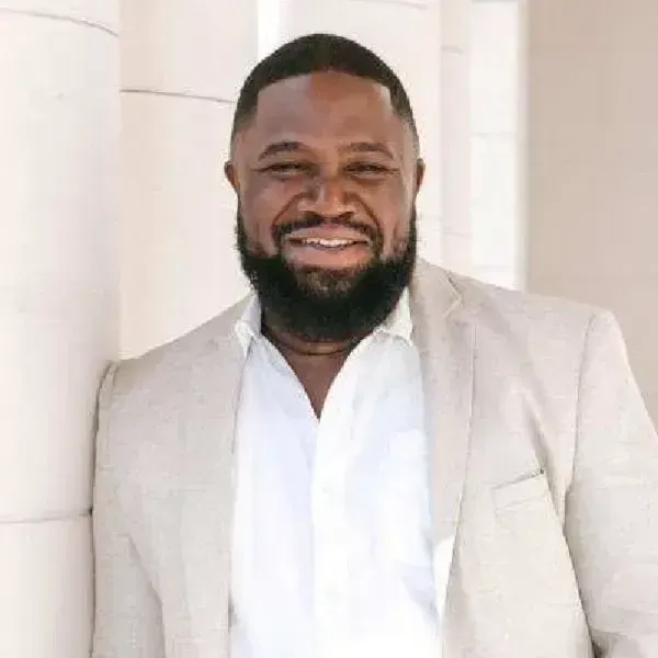 Maurice A. Shabazz smiling in a light beige suit and white shirt standing against a white columned wall.