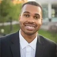 Darrin Harvey smiling in a black suit and white shirt standing outdoors with a blurred building in the background.