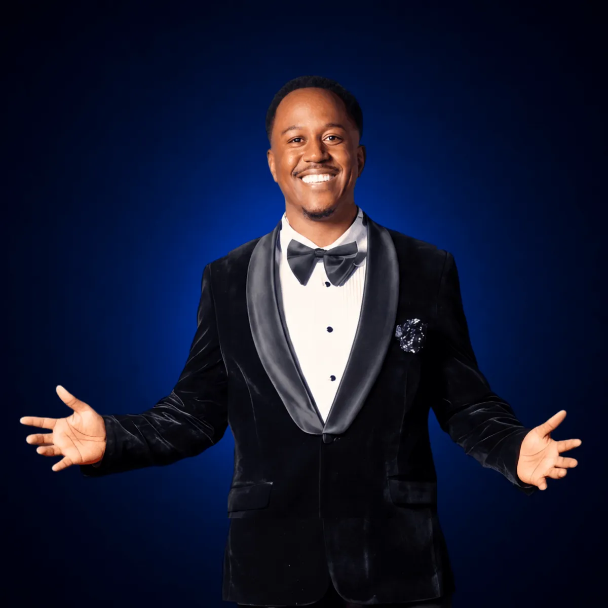 Troy Sandidge smiling in a tuxedo and white shirt posing against a gradient background