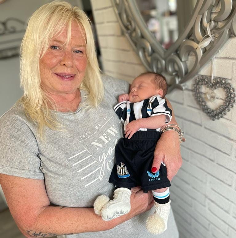 Elaine Somerville with blonde hair, holding her granddaughter Ruby wearing newcastle united football strip, against a grey brick wall, with a silver heart shaped mirror