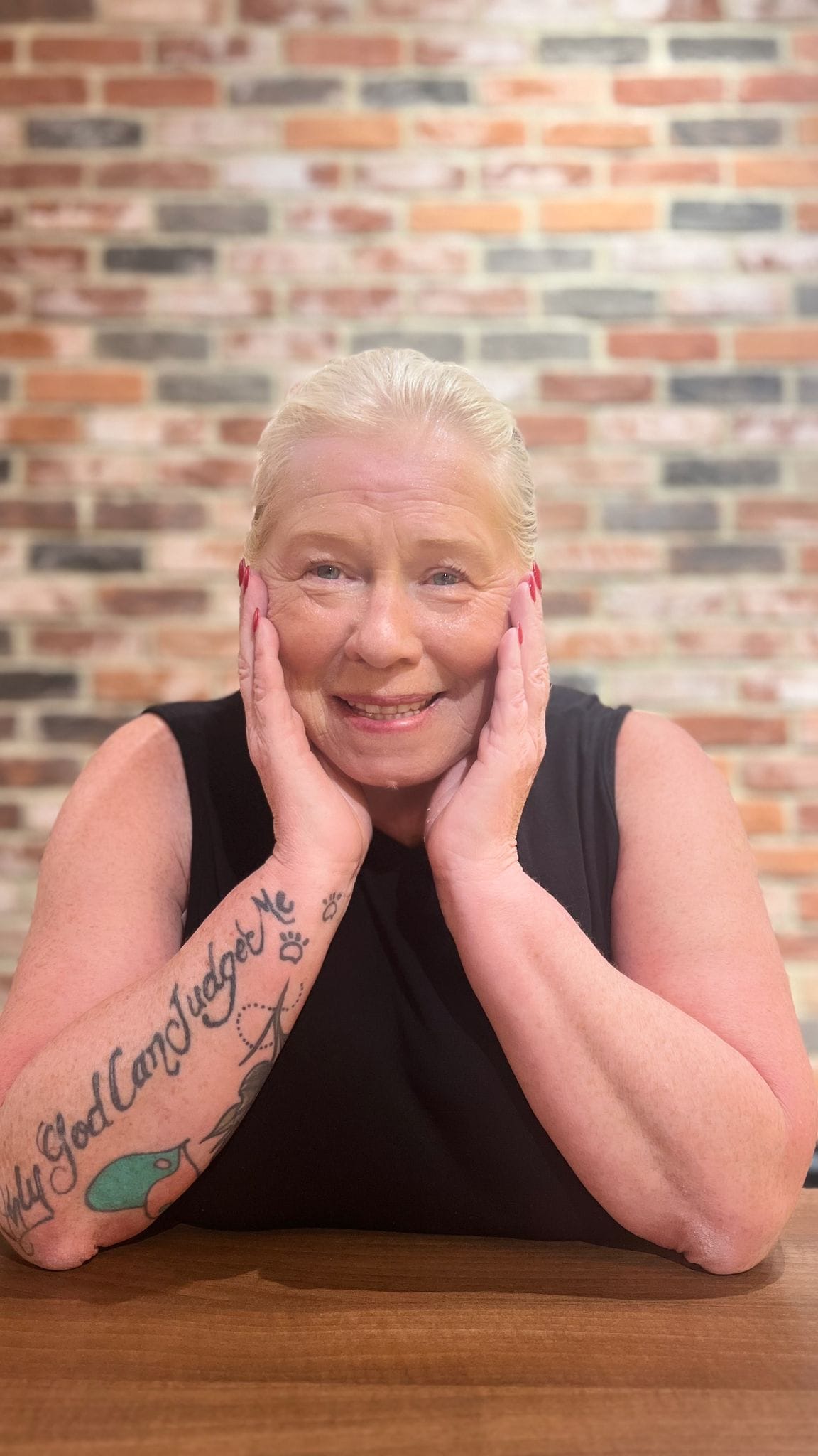 Elaine Somerville with blonde hair, red nails, black top against a brickwall, smiling blue eyes