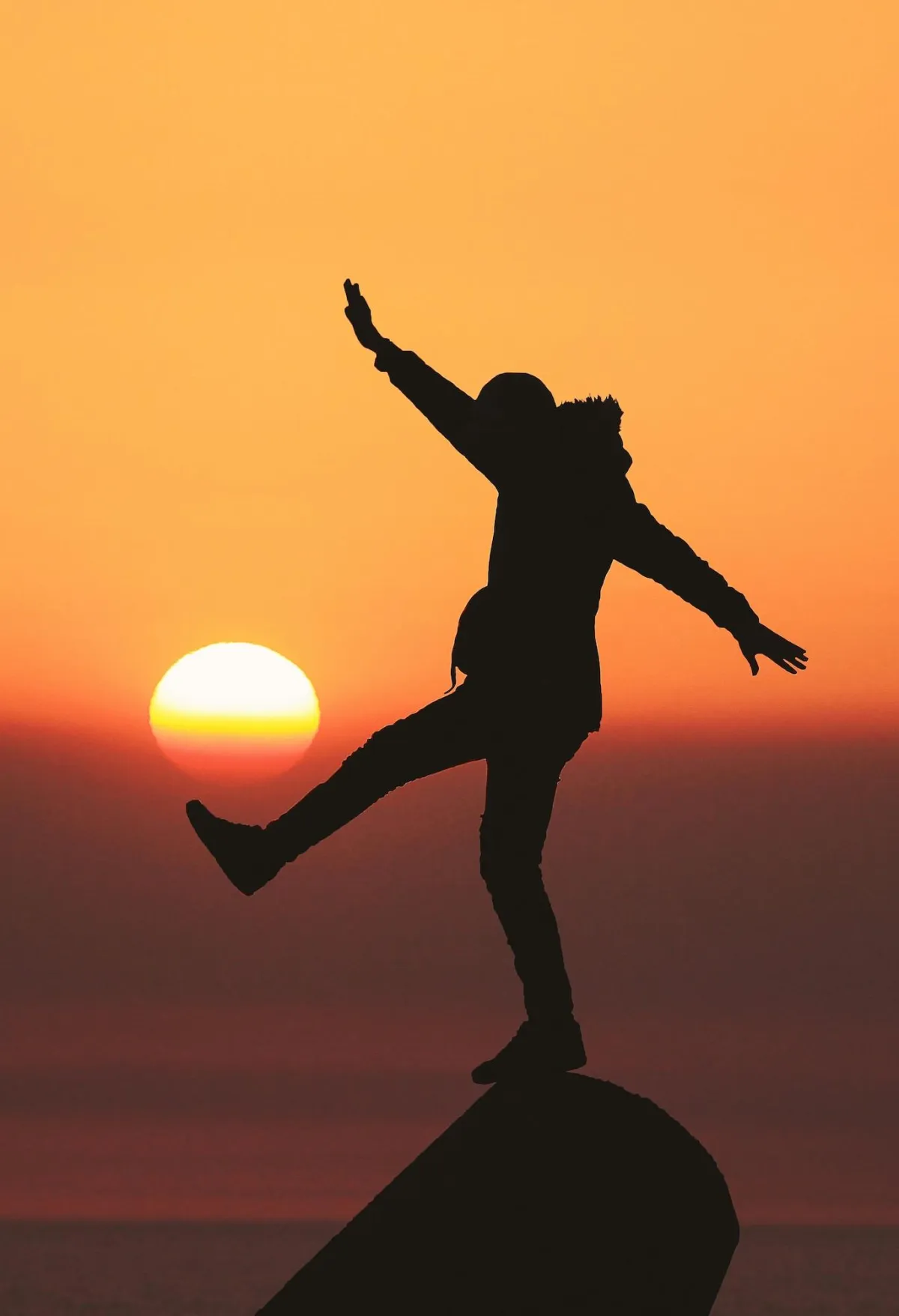 Person balanciert auf einem Felsen bei Sonnenuntergang und es wirkt so als ob die Person mit der Sonne Fussball spielt