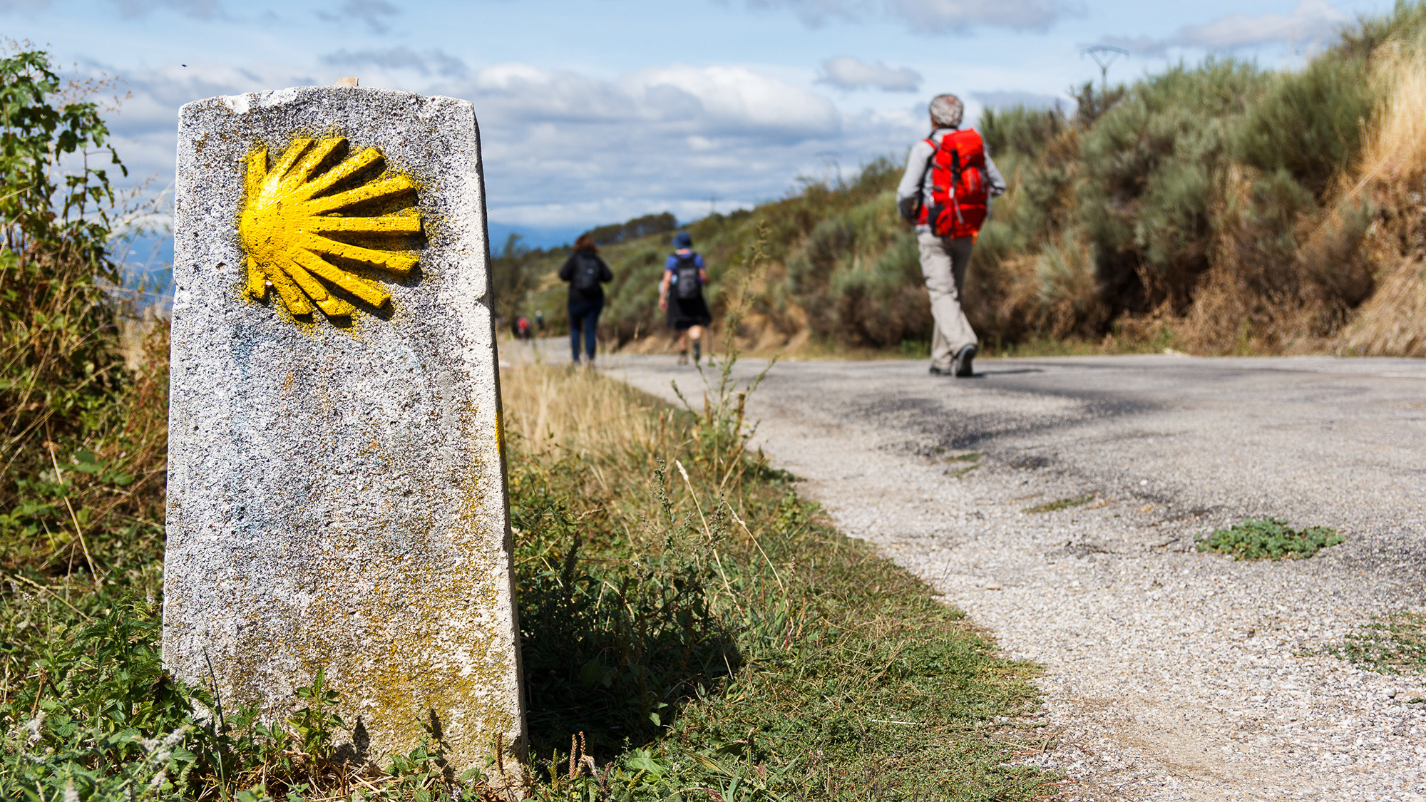 Stone Camino waymarker beside a rural road with pilgrims walking ahead