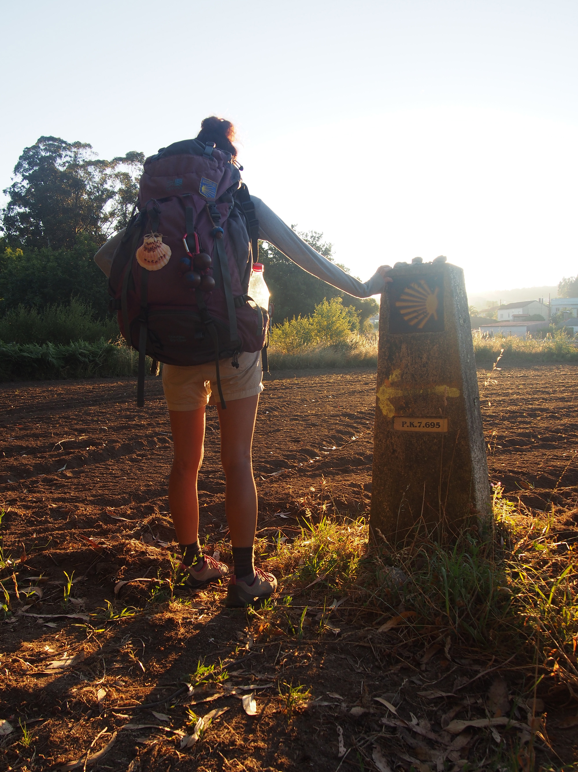 Pilgrim with backpack pausing beside a Camino waymarker in early morning light