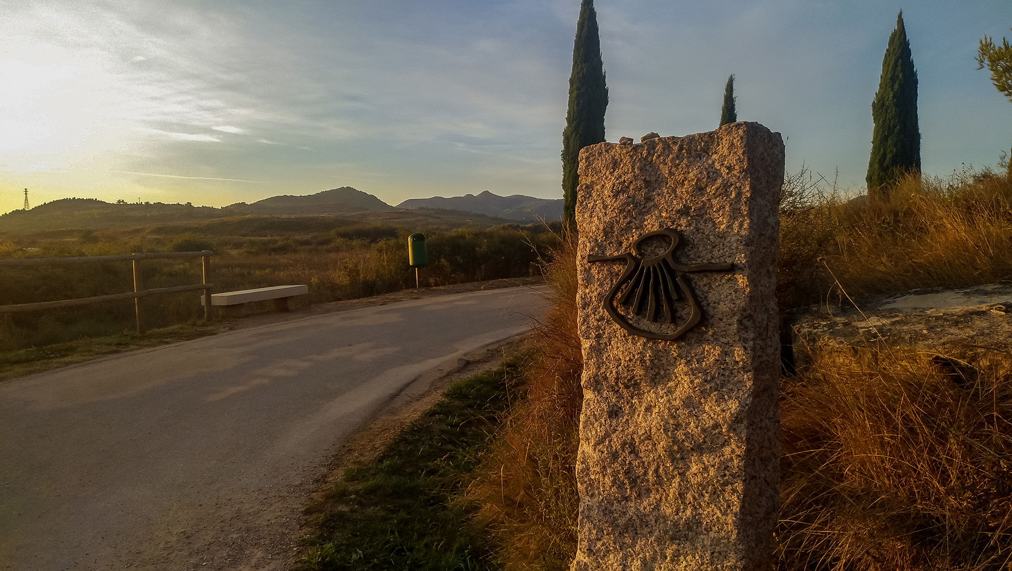 Stone Camino waymarker beside a rural road with hills beyond at sunset