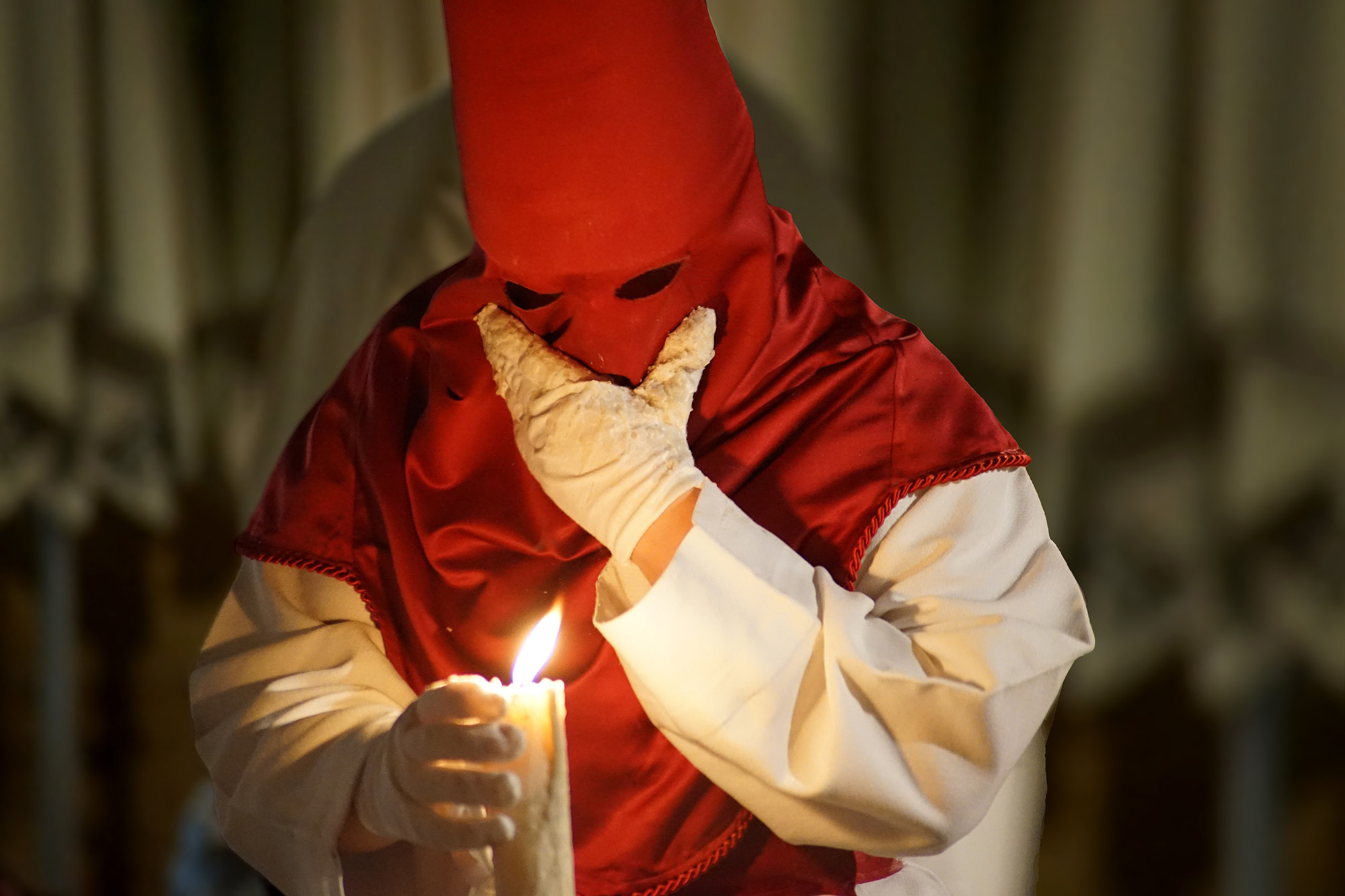 Penitent in red hood and white robe holding a candle during a Semana Santa procession