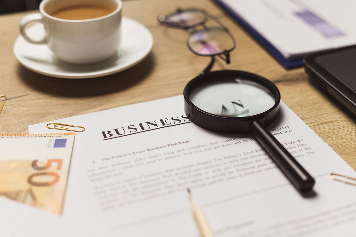 Printed business plan on a wooden desk beside a magnifying glass, reading glasses, coffee and a 50-euro note