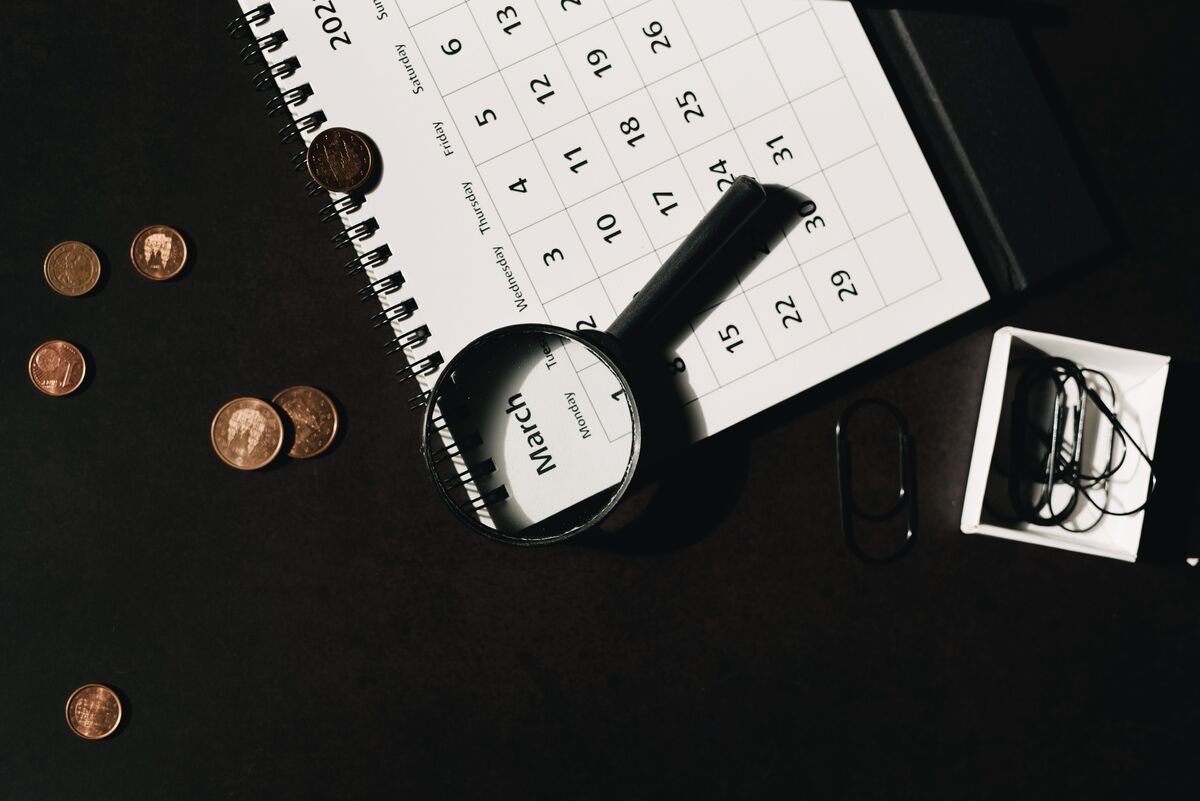 Monthly wall calendar beside scattered coins and a magnifying glass on a dark desk, suggesting months-ahead budgeting