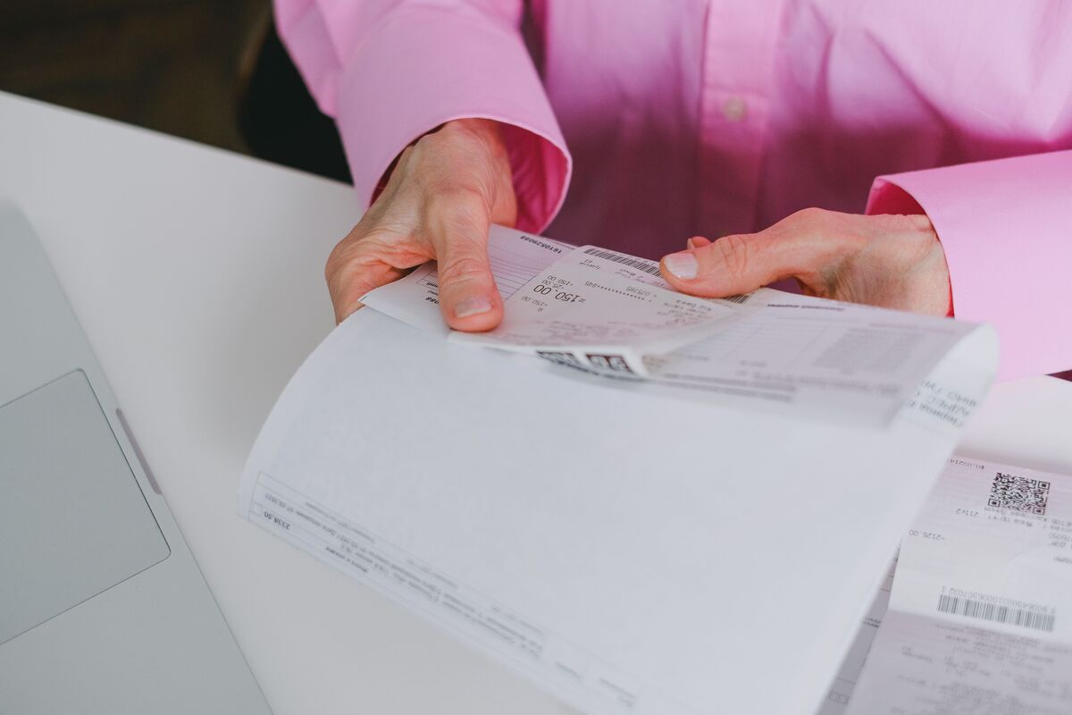 Hands in a pink shirt sorting through paper invoices and receipts at a desk beside a laptop