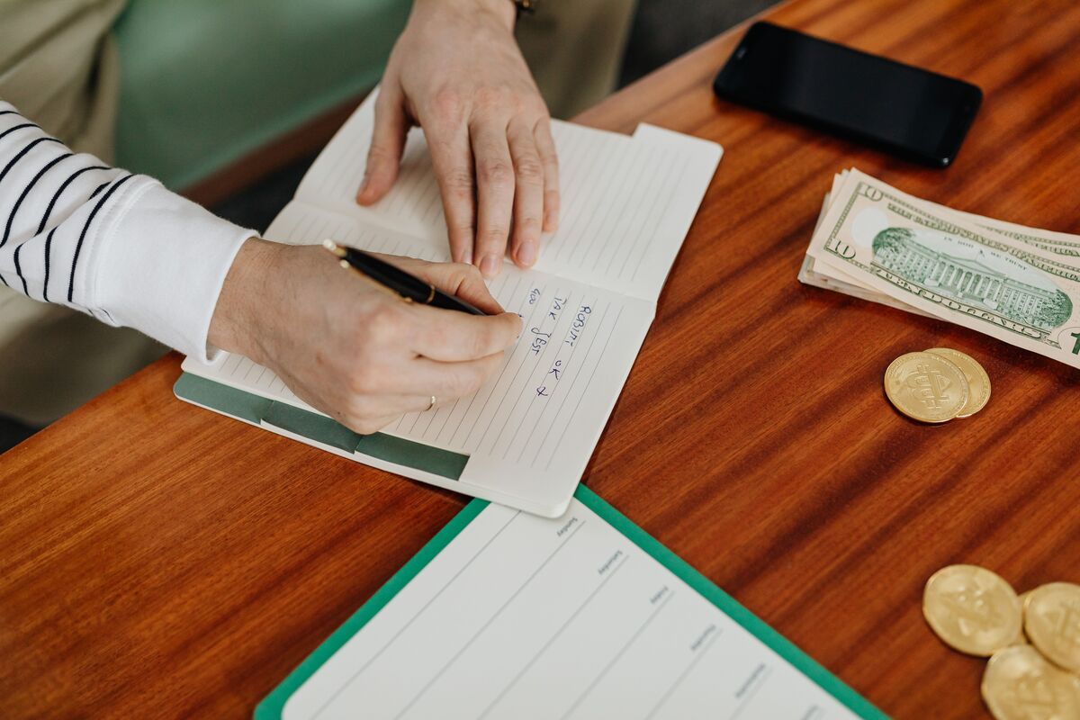 Person writing a budget by hand in a notebook with cash and coins on a wooden desk
