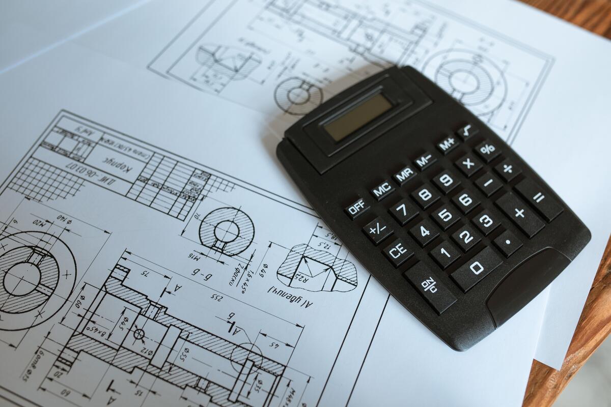 Black calculator resting on detailed engineering blueprints with technical drawings on a wooden desk
