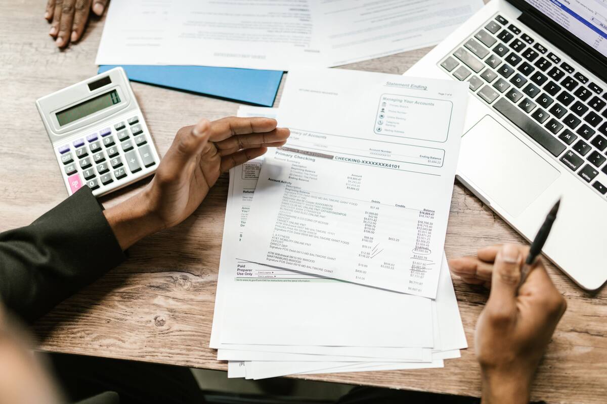 Person reviewing a bank statement beside a calculator, laptop and pen on a wooden desk during account reconciliation