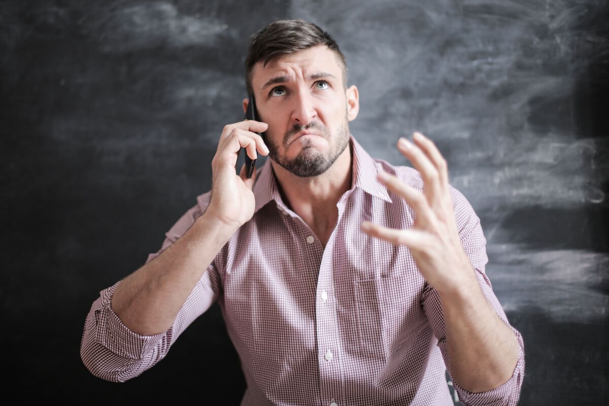 Worried man in a checked shirt frowning during a phone call against a dark wall, one hand raised in frustration