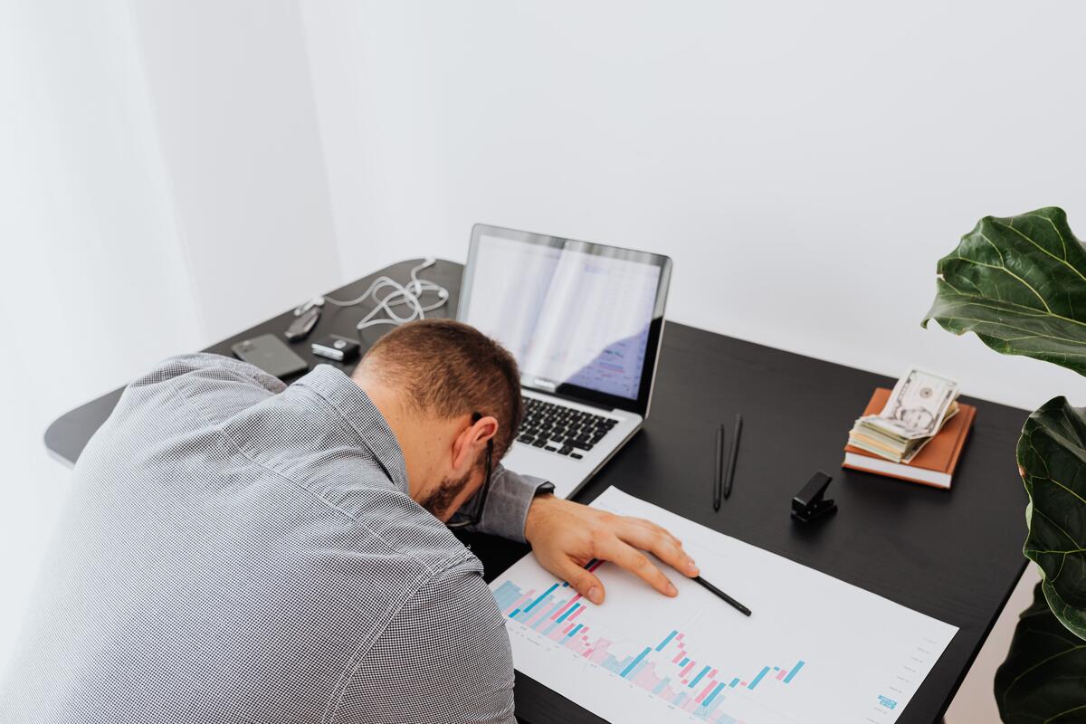 Exhausted business owner slumped at his desk over a financial bar chart, laptop and cash nearby