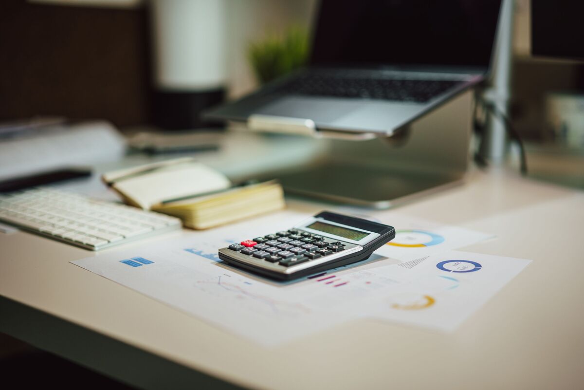Calculator resting on colorful financial charts beside a laptop and notepad on a clean, bright office desk