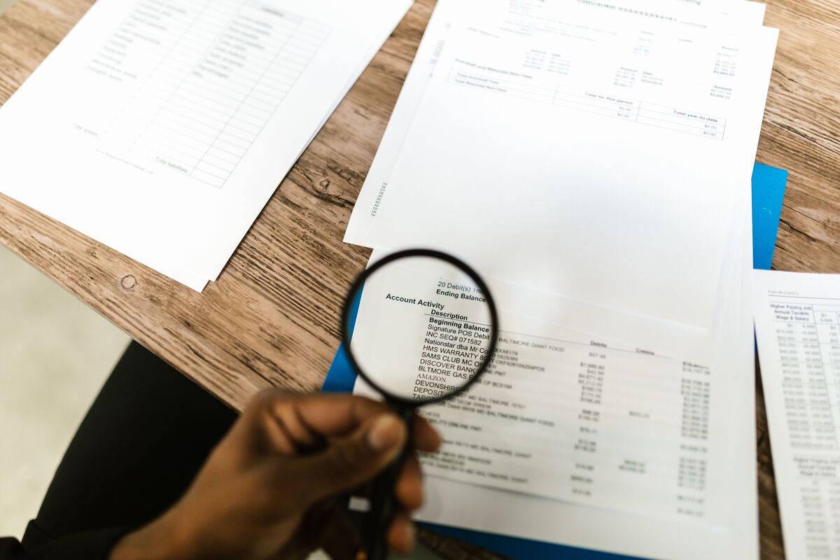 Hand holding a magnifying glass over financial statements and account records spread across a wooden desk
