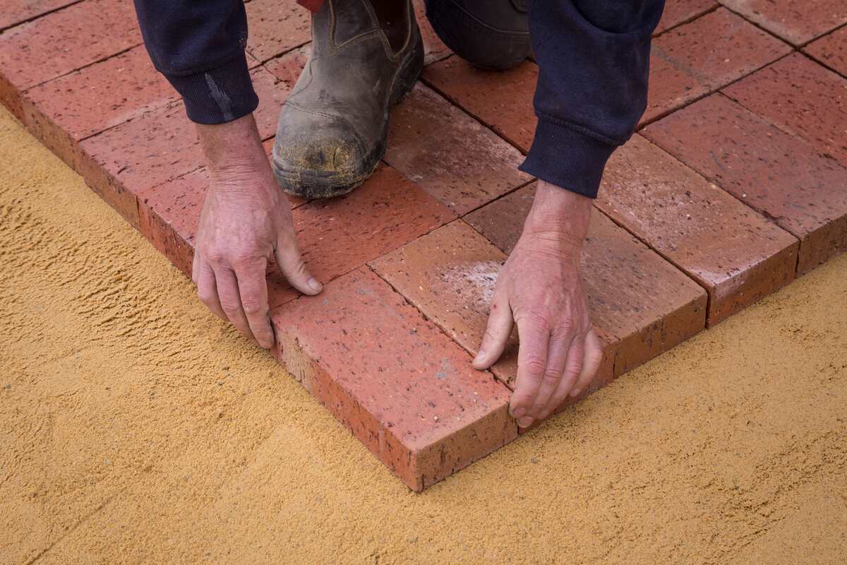 Worker's hands laying red clay pavers on a sand bed while installing a patio corner