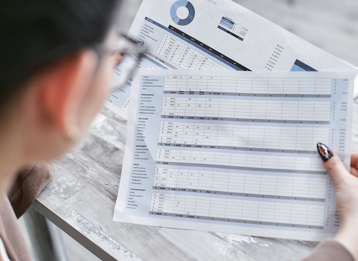 Person reviewing a printed budget spreadsheet with monthly columns, line-item totals and a pie chart at a desk