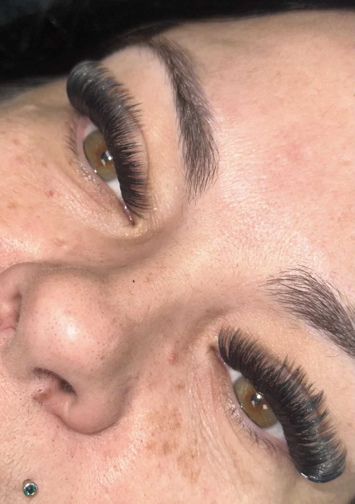 A client with a deep skin tone and closed eyes showcases lush, voluminous lash extensions. The image is tightly cropped, with a soft ivory background and subtle gold lighting for a luxurious, editorial feel.