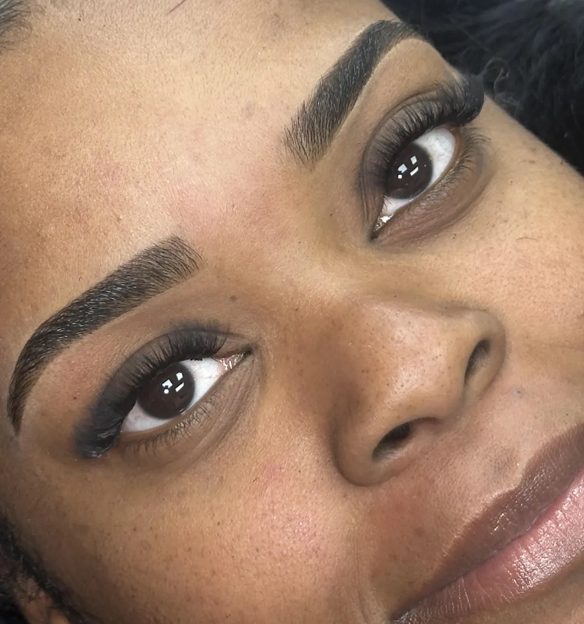 A close-up of a client's face highlighting expertly shaped brows, with natural skin texture and a gentle smile. The background is softly blurred in beige tones, emphasizing the precision and artistry of the brow work.