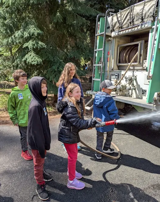 Students hiking a nearby trail under late-afternoon light with backpacks and a teacher guide, forested foothills of the Cascades visible, photorealistic action shot showing outdoor learning, exploration, and community.