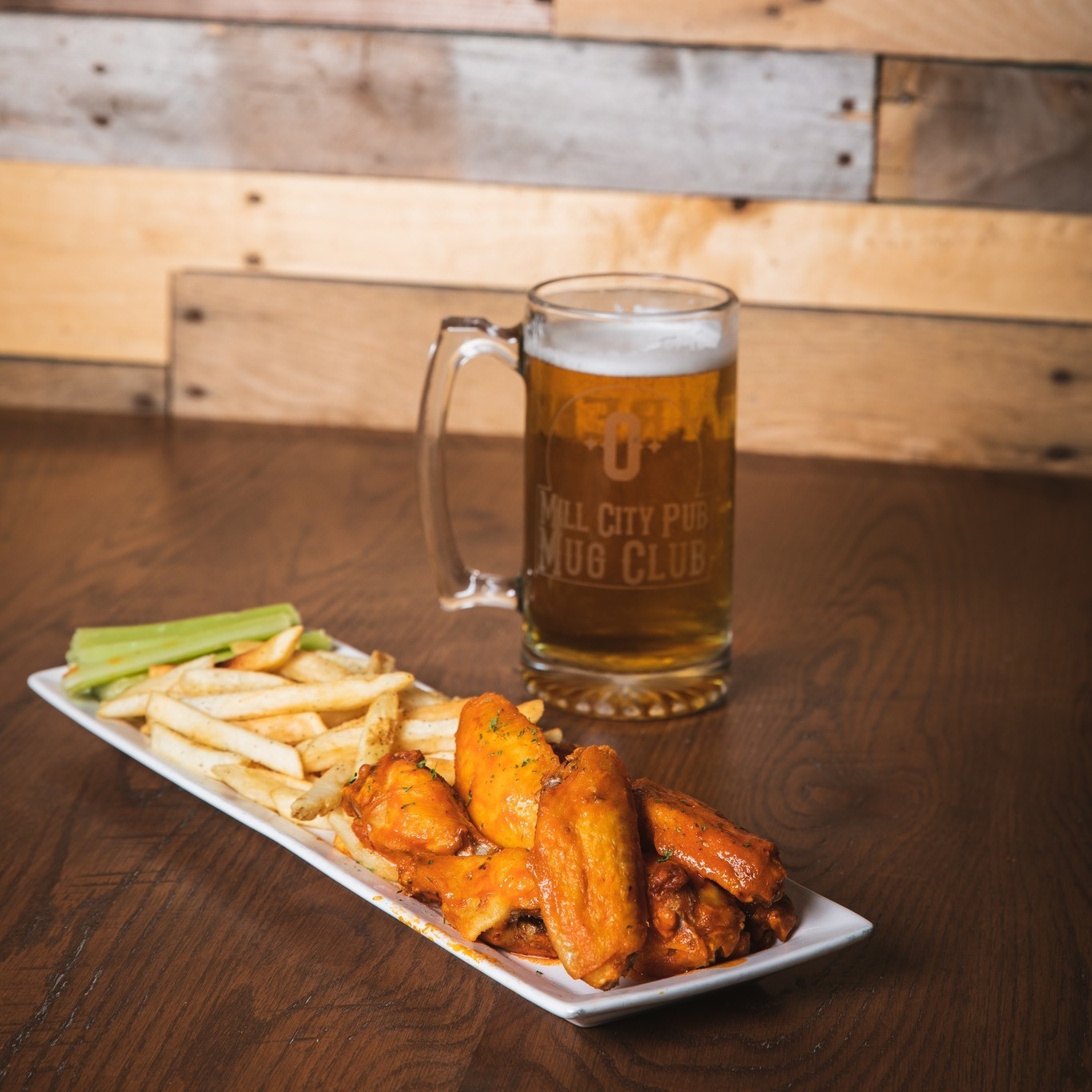 A plate of buffalo wings and a side of fries sitting next to a mug filled with light beer