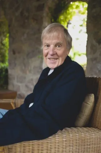Bob Frissell seated in a warm study, medium shot 1:1 portrait format, natural window light, books and plants blurred in background, relaxed pose, candid photorealistic moment conveying approachability and lived experience.