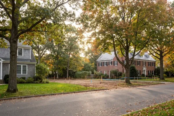 A tree-lined North Shore street with a shingle-style house and a brick Georgian-revival house beside an empty lot staked out with ribbon