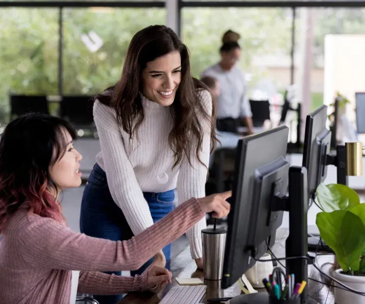 Two women collaborating at a desk with computers, one pointing at the screen, in a modern office setting, reflecting teamwork and IT support solutions.