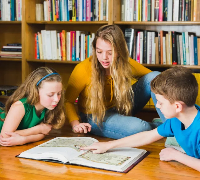 A smiling female tutor and a young girl work together at a desk in a classroom. The girl is filling out a colorful graphic organizer with a pencil while the tutor observes supportively
