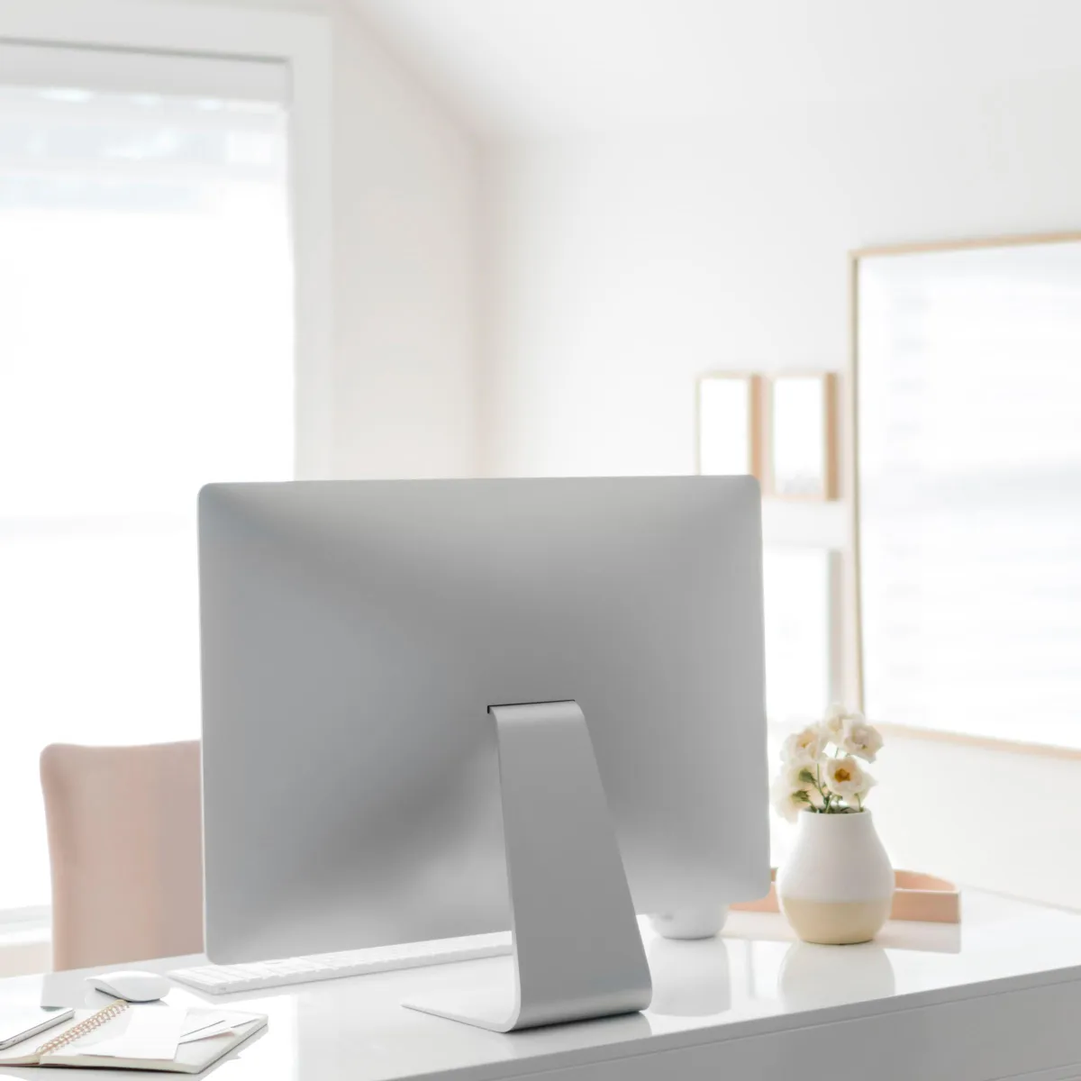Back side of a silver computer monitor on a white desk with a small flower vase.