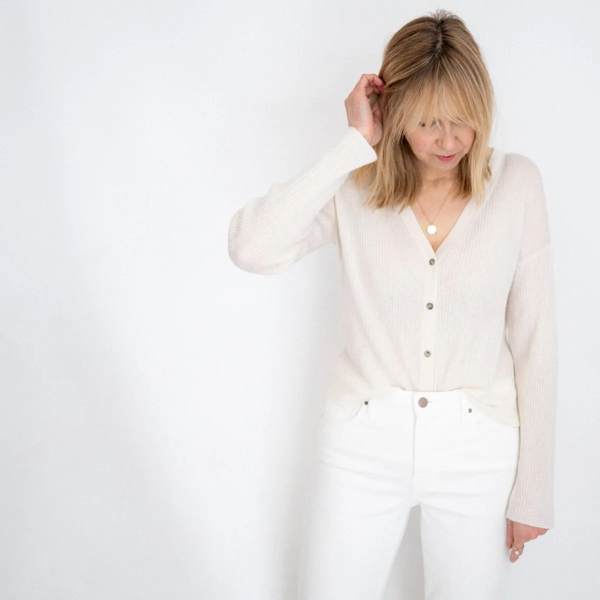 Mature woman standing in front of a white wall looking down with straight face.