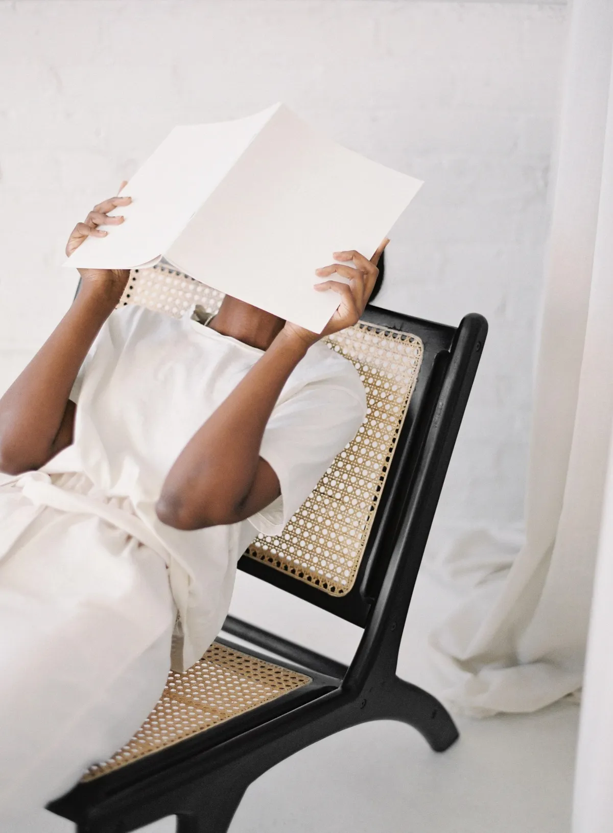 Woman in a chair holding a folder over her face, seemingly frustrated.